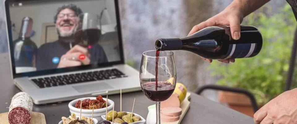 Person pouring red wine into wine glass having virtual drinks on laptop with various snacks on table