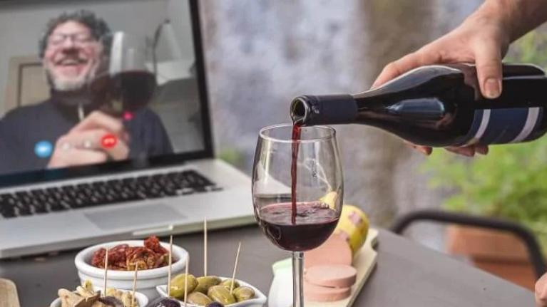 Person pouring red wine into wine glass having virtual drinks on laptop with various snacks on table