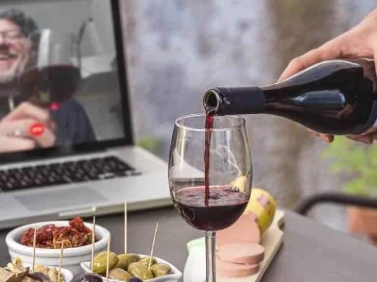 Person pouring red wine into wine glass having virtual drinks on laptop with various snacks on table